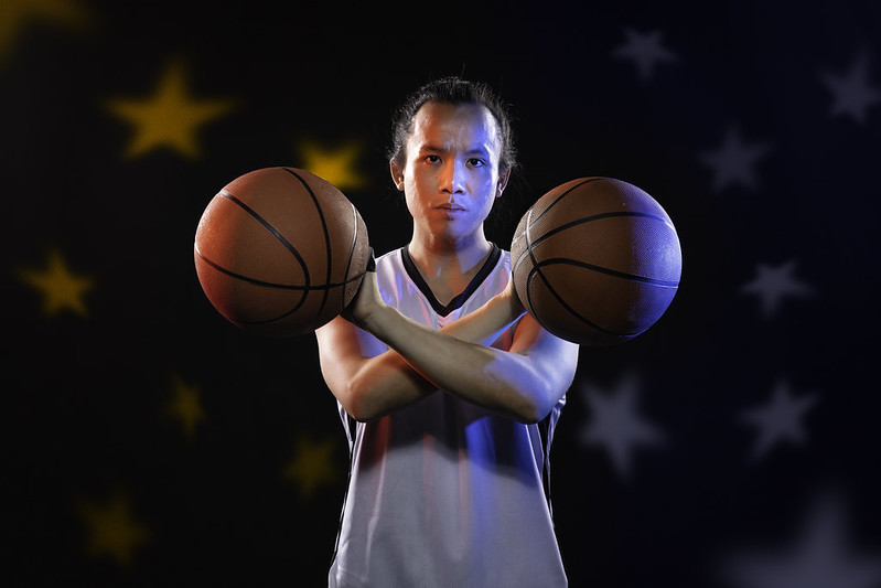 A young man palms two basketballs crossed across his chest; red and blue lights illuminate him from either side