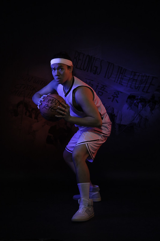 A young man crouches and holds a basketball; images of the Tiananmen Square Protests are illuminated in red and blue light