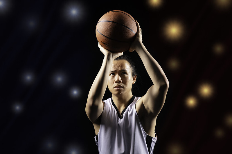 A young man holding a basketball focuses intensely as he makes a free throw; camera flashes are seen behind him