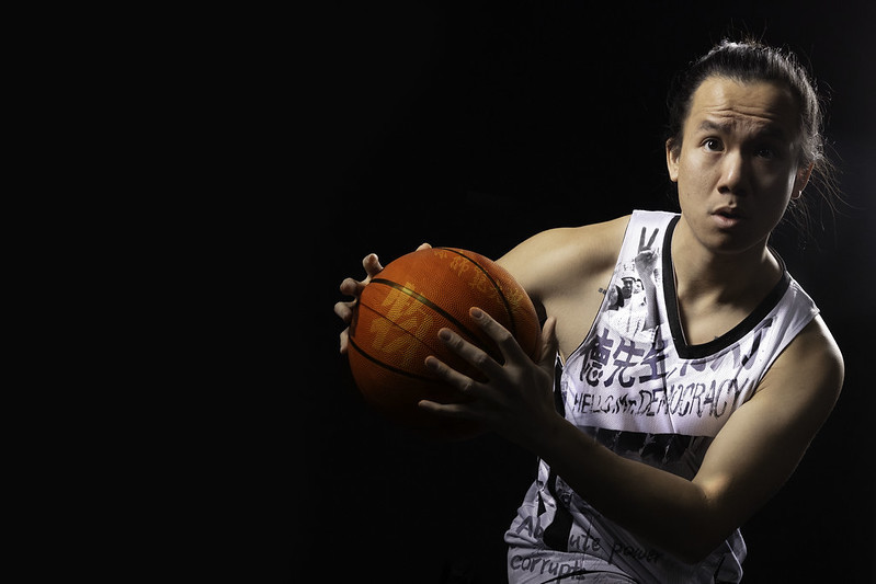 A young man with a concerned look holds out a basketball; images of the Tiananmen Square Protests are faded across his jersey