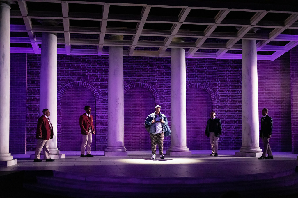 Among columns and brick archways a Black man in street clothes is flanked by four other young Black men in school uniforms.