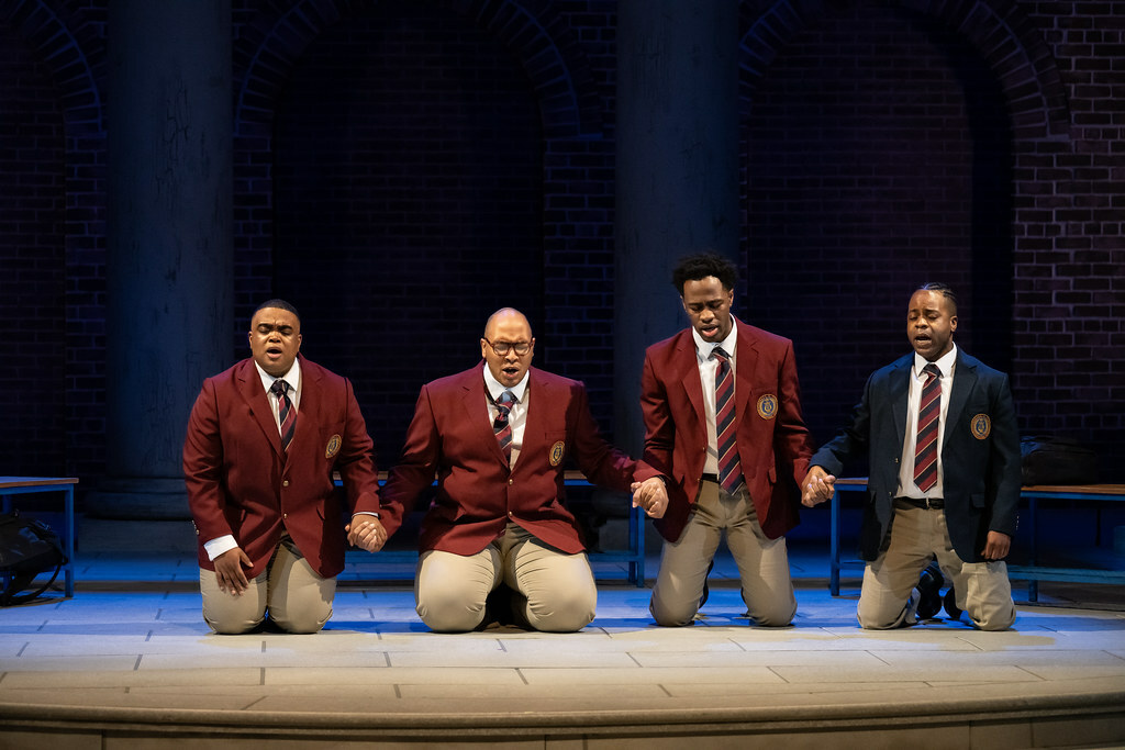 Four young Black men in school uniforms kneel on the ground together, holding hands.