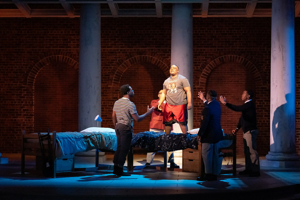 A young Black man in a t-shirt and athletic shorts stands on a bed as four others surround the bed, arms raised toward him.