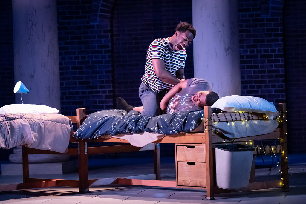 Two young Black men are roughhousing and laughing on a dormitory bed.