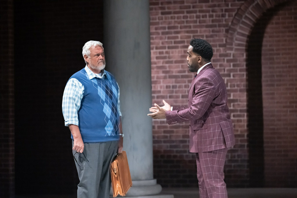 A Black man in a suit gestures fervently, talking to an older white-haired white man in a sweater vest, holding a briefcase.