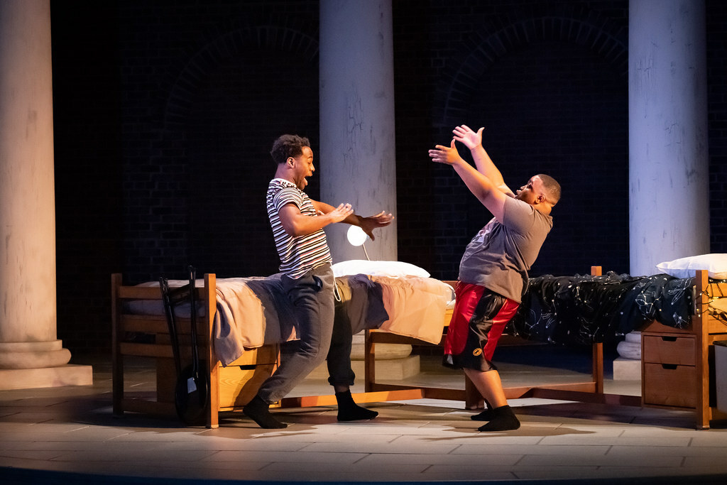 Two young Black men in t-shirts, sweats/shorts, and socks gesture excitedly to each other in front of two dormitory beds.