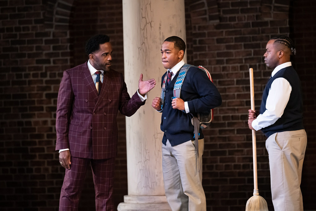 A Black man in a suit gestures, talking to two young Black men in school uniforms, one wearing a backpack, one with a broom.