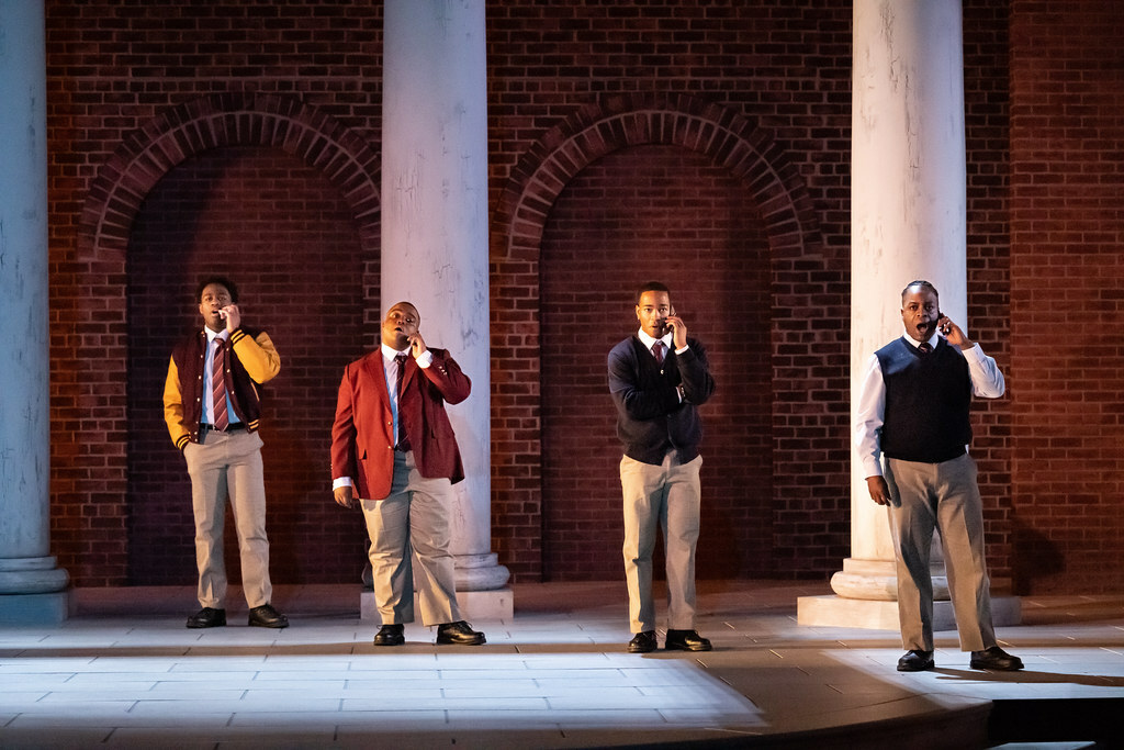 Four young Black men in school uniforms stand before marble columns, singing with cell phones held to their ears.