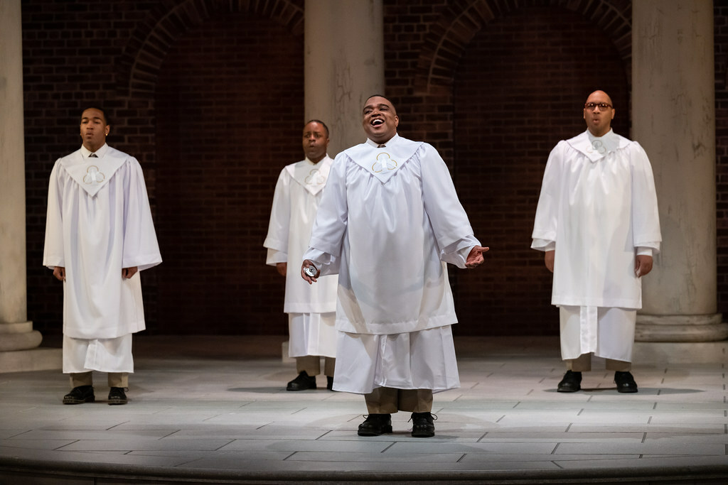 Four young Black men in white church robes sing while standing in front of marble columns.