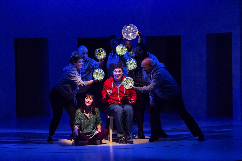 A young man and woman sit in a spotlight surrounded by glowing orbs 