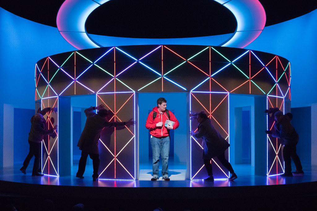 A young man stands center, surrounded by colorful LED lights and bodies reaching out towards him