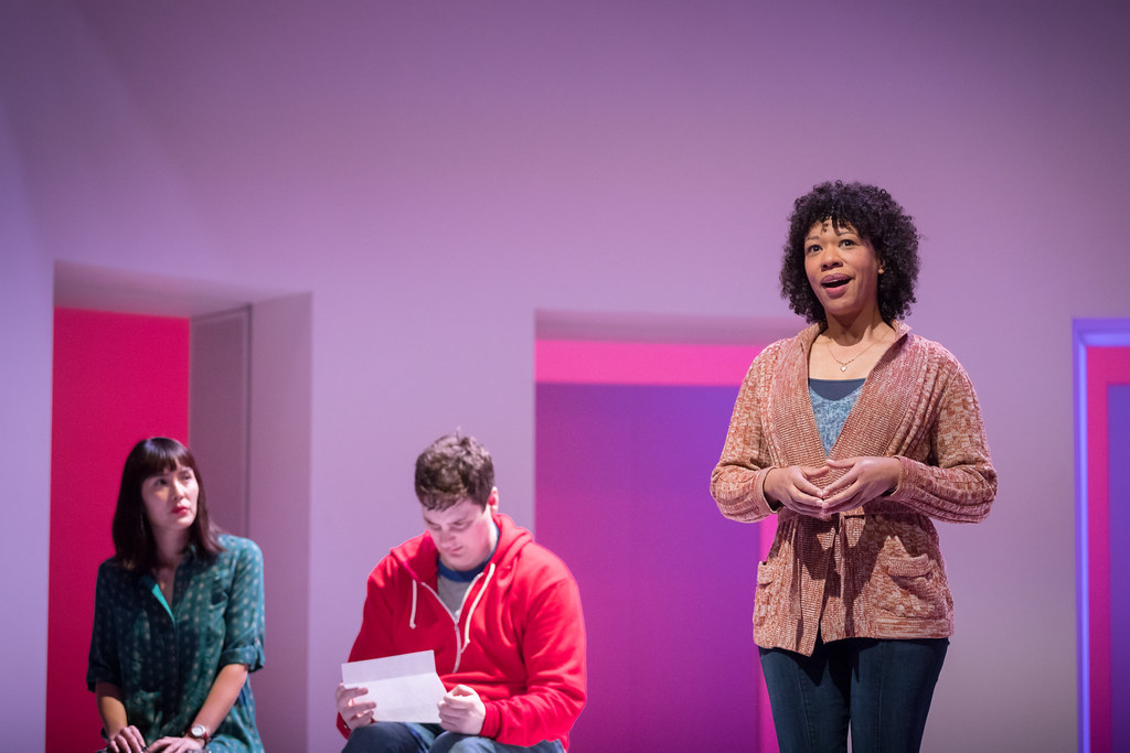 A woman stands in the foreground smiling while a man and woman sit behind her intently focused