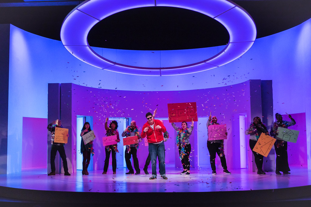 A young man is showered in confetti surrounded by a joyful group of people holding colorful signs