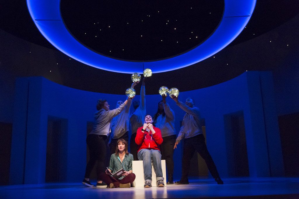 A man seated on a block, onstage in a spotlight, with a woman seated on the floor next to him holding a book. People them hol