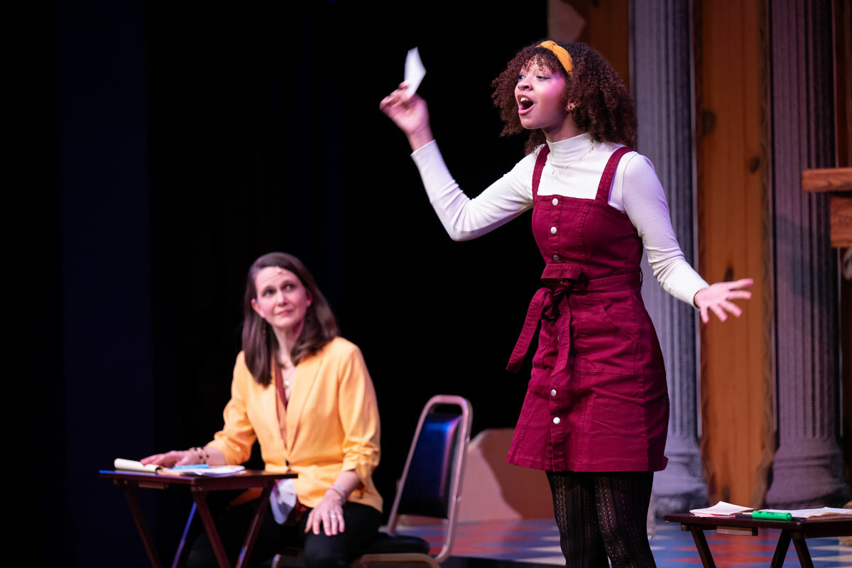 A young woman stands speaking demonstratively, holding up a paper in one hand, as an older woman seated at a small writing desk looks on.