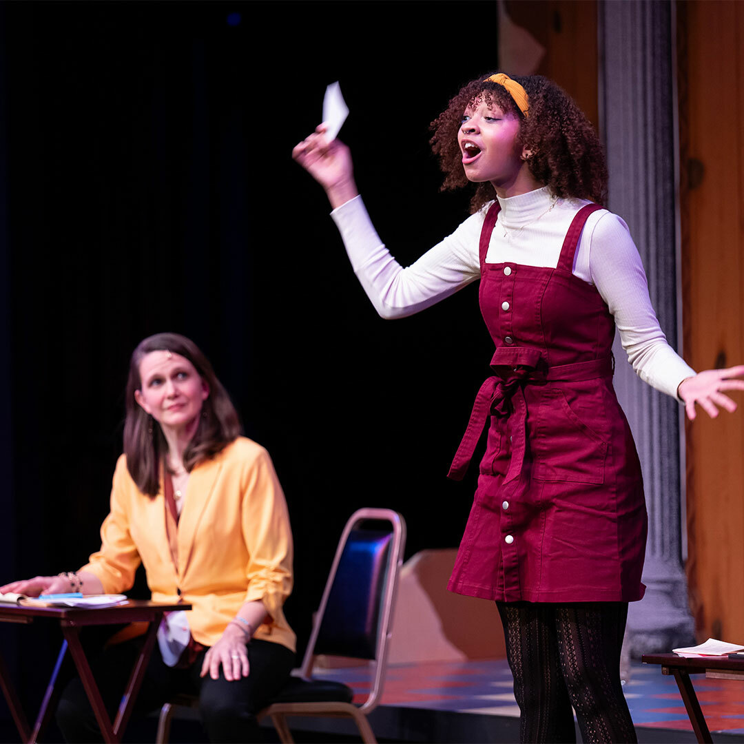 A young woman stands speaking demonstratively, holding up a paper in one hand, as an older woman seated at a small writing desk looks on.