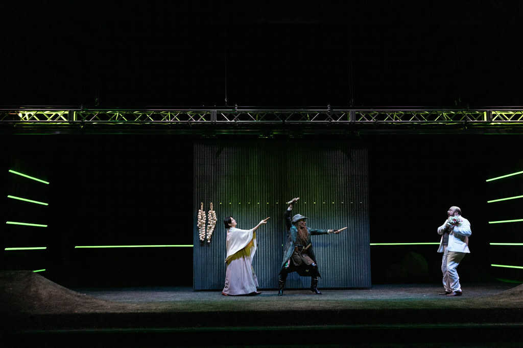 On a barren stage lit with narrow bands of eerie green light, two women brandish sharp wooden stakes at a cowering man.