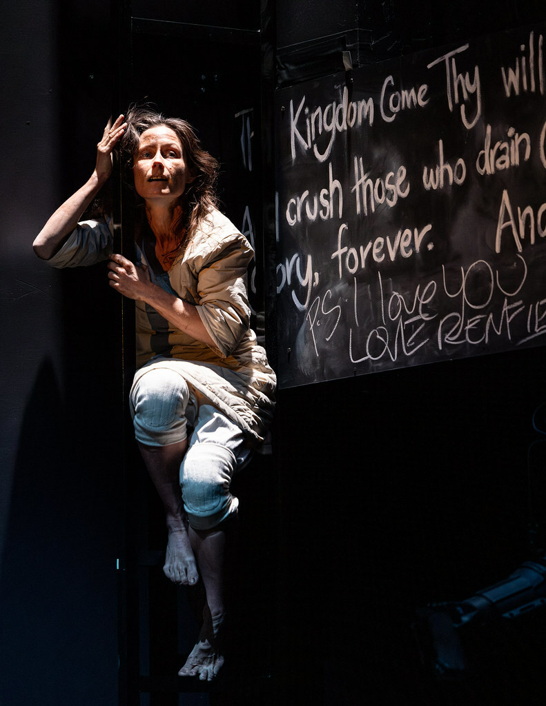 A distressed woman in pale, ragged clothes and bare feet perches next to a black panel covered with scribbled white text.