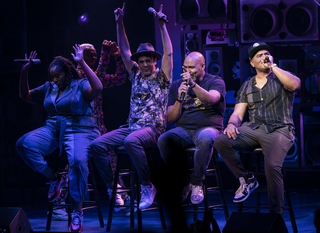 Five performers are onstage; several raise their hands in hype while several others are mid-speech