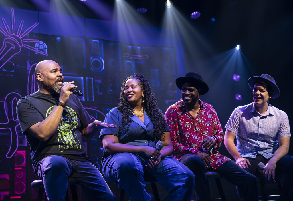 Four performers sit on stools; three look off to their right smiling at the fourth who is mid-speech