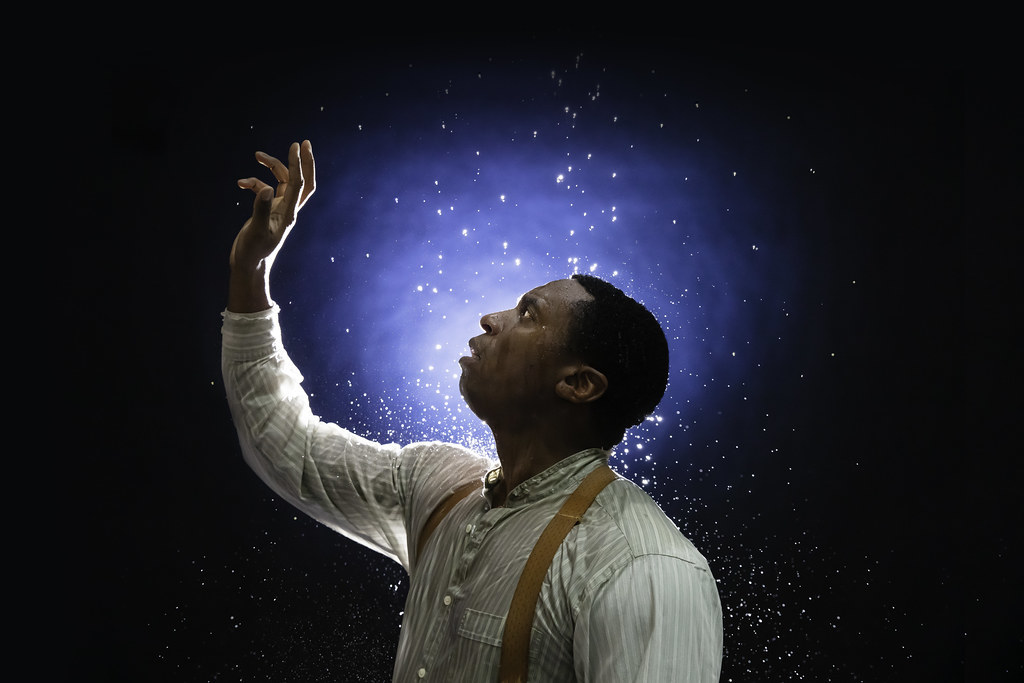 A young man gazes at his hand, water droplets suspend in the air around him; a blue background illuminates him from behind
