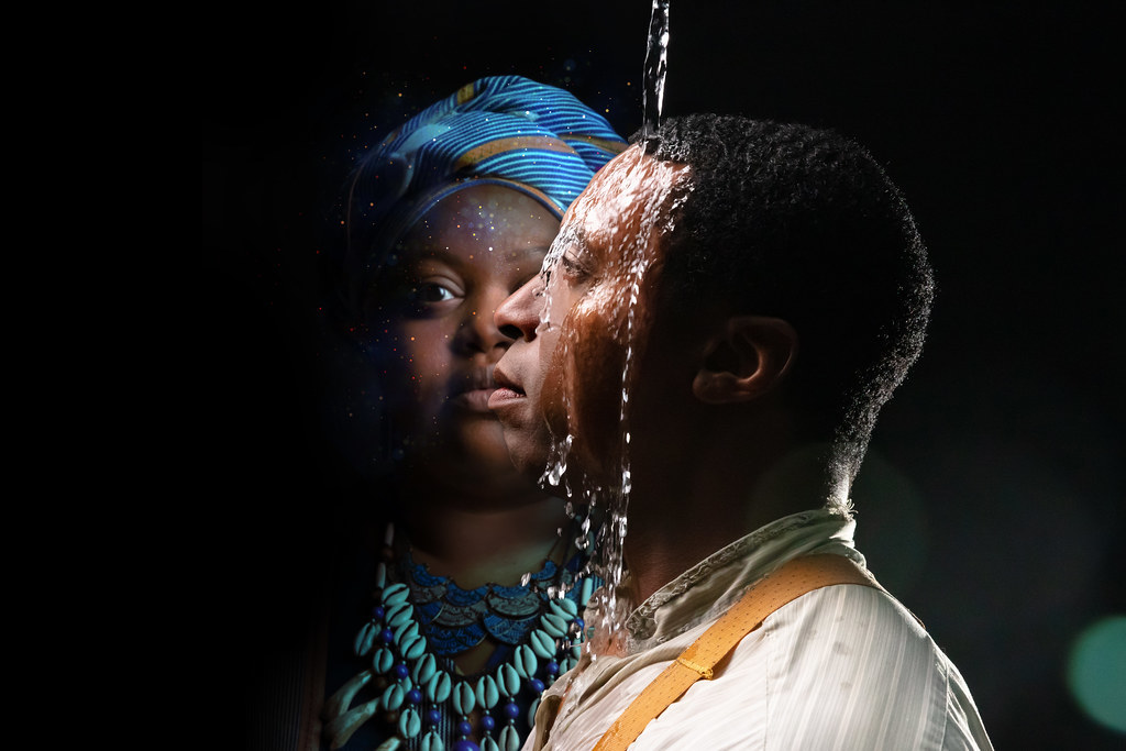 A man stands in the foreground as water falls on his head; another person stands staring forward in the foreground