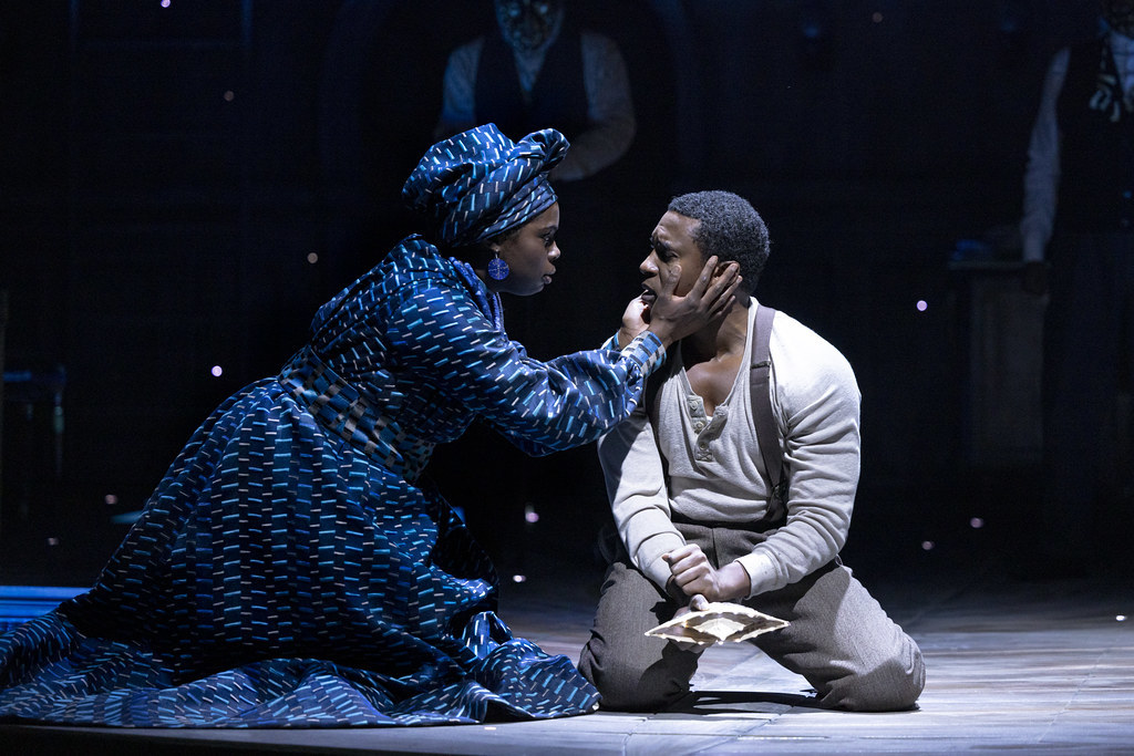 A young woman holds a young man's face between her hands; they are both kneeling on the ground.