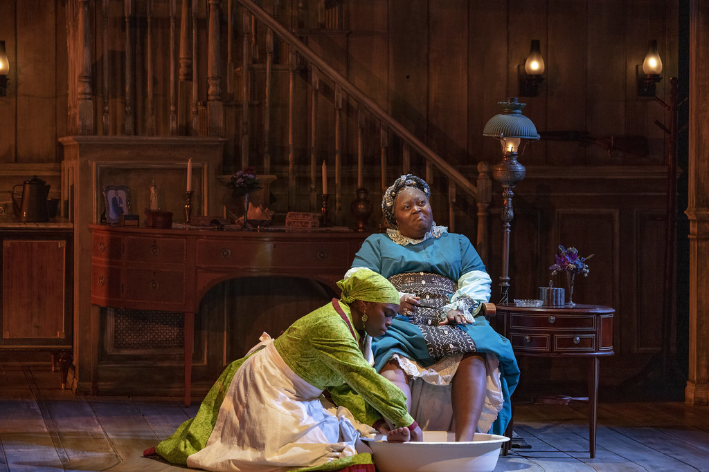 A woman washes a person's feet as they look out, contemplative.