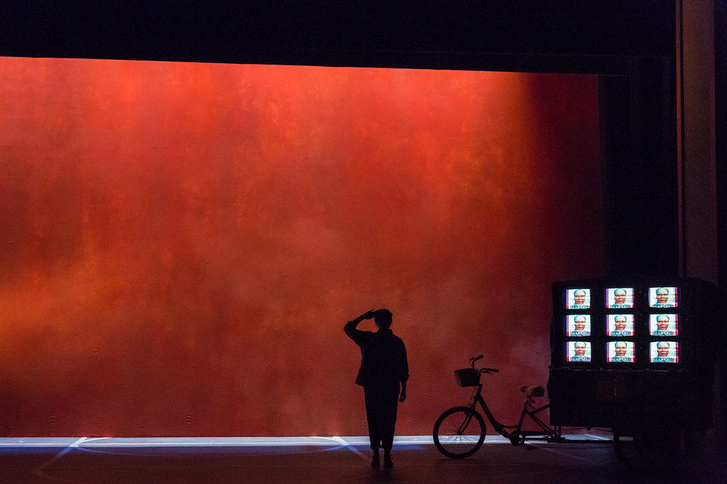 A body is silhouetted against a bright red backdrop; a bicycle-pulled cart holds nine TVs displaying the image of Mao Zedong