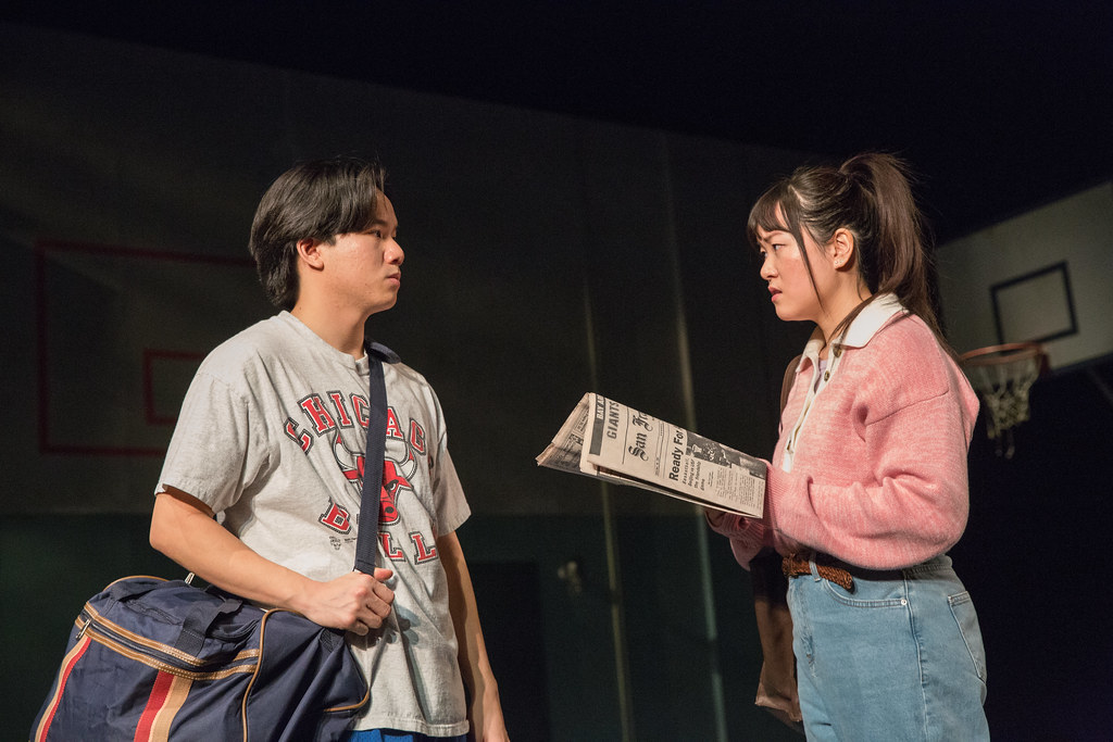 A young man and a young woman are caught in a tense stare; the young woman holds a newspaper