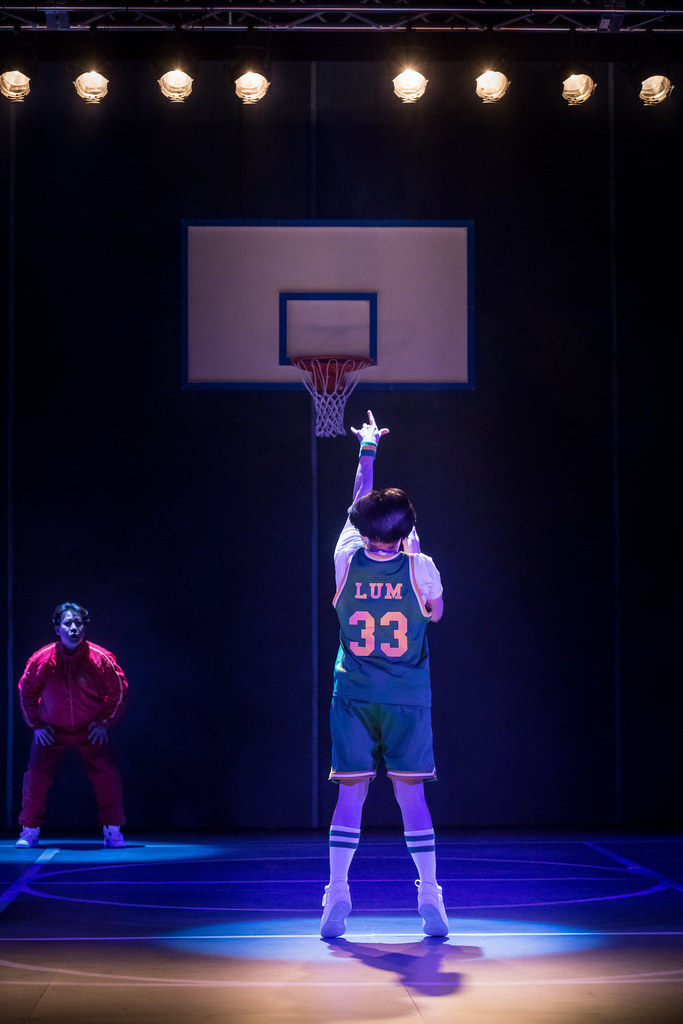 A man watches a younger man take a free throw; they are frozen in a tense, dramatic moment