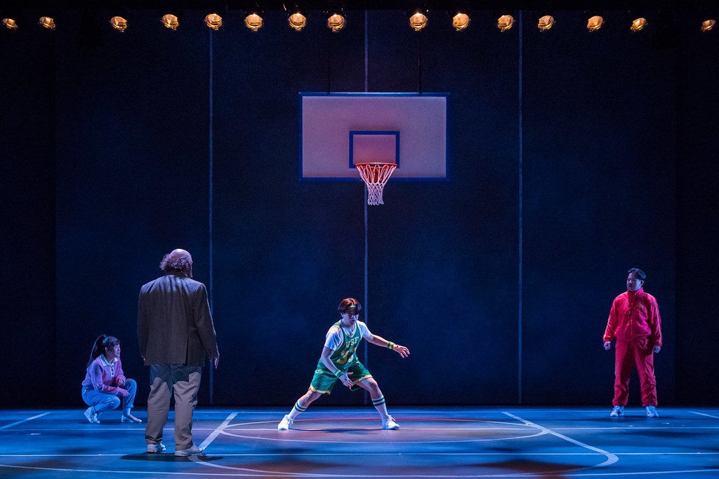 Three people on a basketball court watch a younger man pantomiming dribbling a basketball intensely