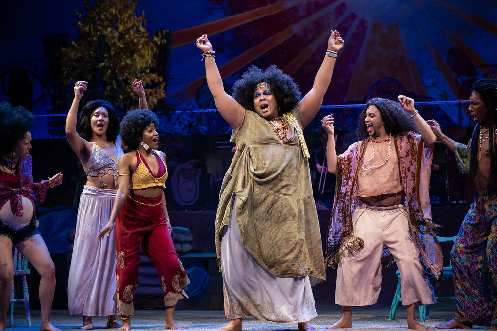 Several young bohemian people of color dance and sing with arms raised.