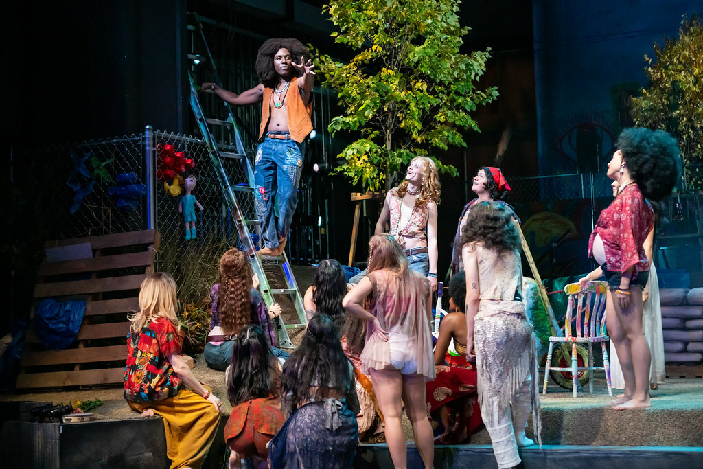 Several young bohemian people look on as a young black man in jeans and a vest stands on a ladder gesturing dramatically.