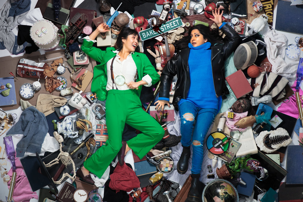 Two women lay atop a pile of curios, trash, and knick knacks. They look at each other, one smiling and the other in distress.