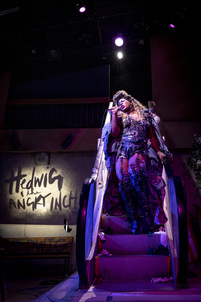 Hedwig strides down an inoperable escalator; the words "Hedwig and the Angry Inch" are projected behind her.