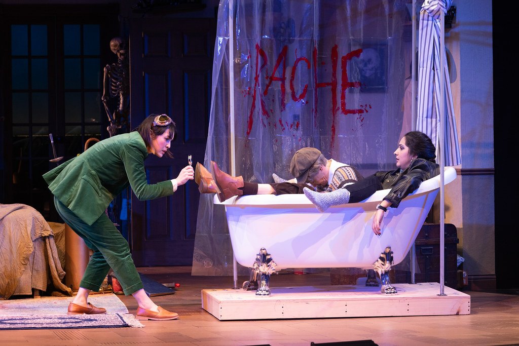 A woman in a bathtub with a dead body, both fully dressed, as another woman checks the body's shoes with a magnifying glass.