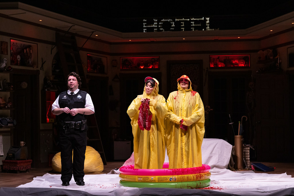 Two women stand in an inflatable kiddie pool wearing yellow rain ponchos dripping with blood. A police officer stands nearby.