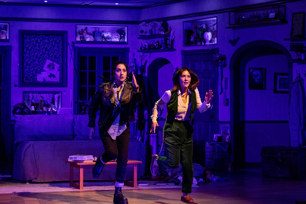 Two women side by side in a cluttered room, facing outward and running in pantomime.