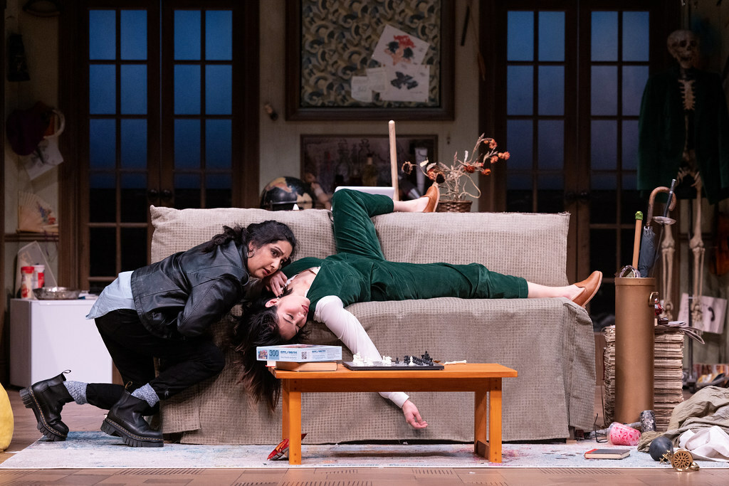 An unconscious woman sprawls awkwardly on a couch, as another woman kneels and checks her neck for a pulse.