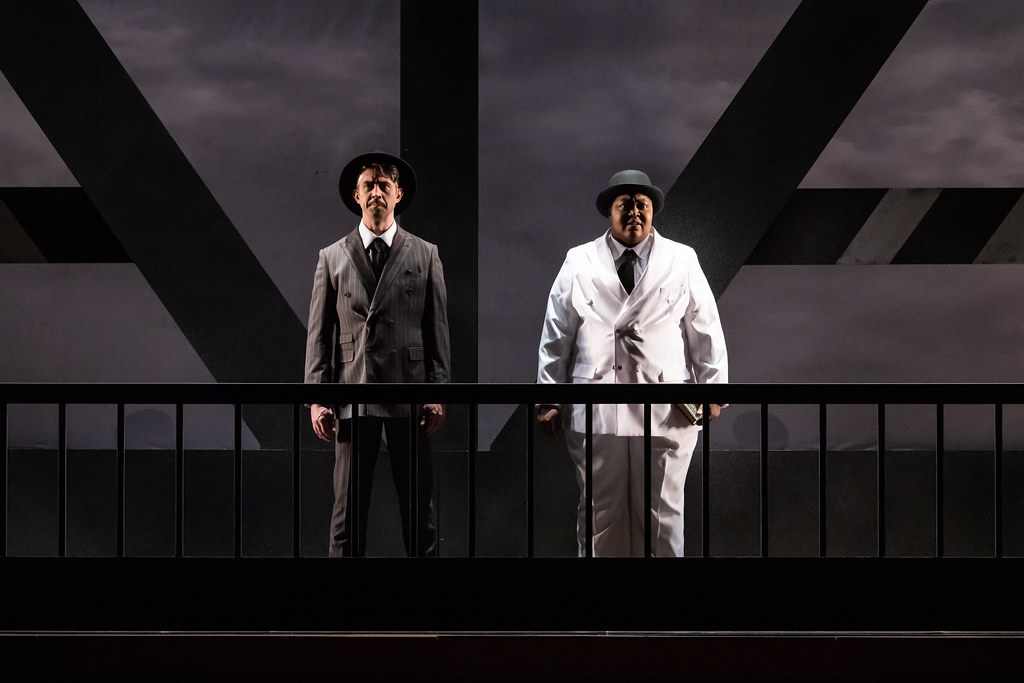 Two people stand in dramatic lighting facing outward on a bridge. The entire stage is black & white including their costumes.