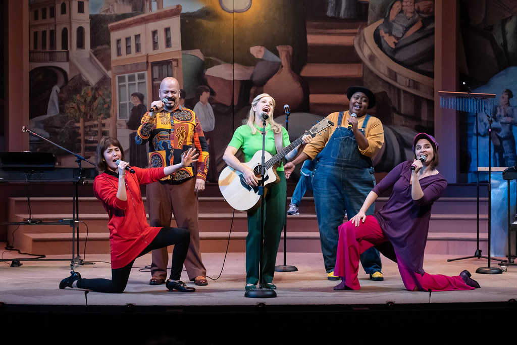 A group of people perform a musical number, posed around a person playing a guitar.