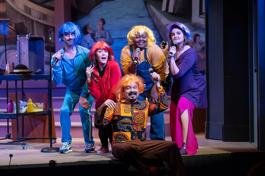 A group of people pose and sing while holding mics. They wear multi colored wigs that match their clothing.