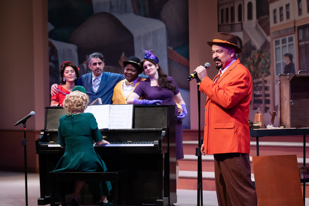 A group of people sing and embrace each other behind a piano. A man in the foreground speaks into a microphone. 