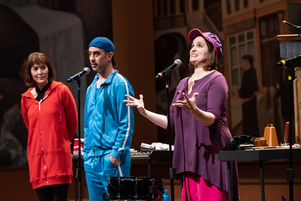 A group of people stand at microphones. A woman in purple spreads her hands upward, her face expressive and big.