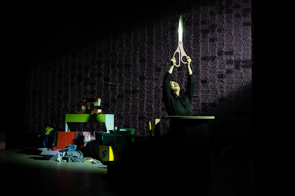 A woman holds up a comically large pair of scissors above her head in a very dramatic light.