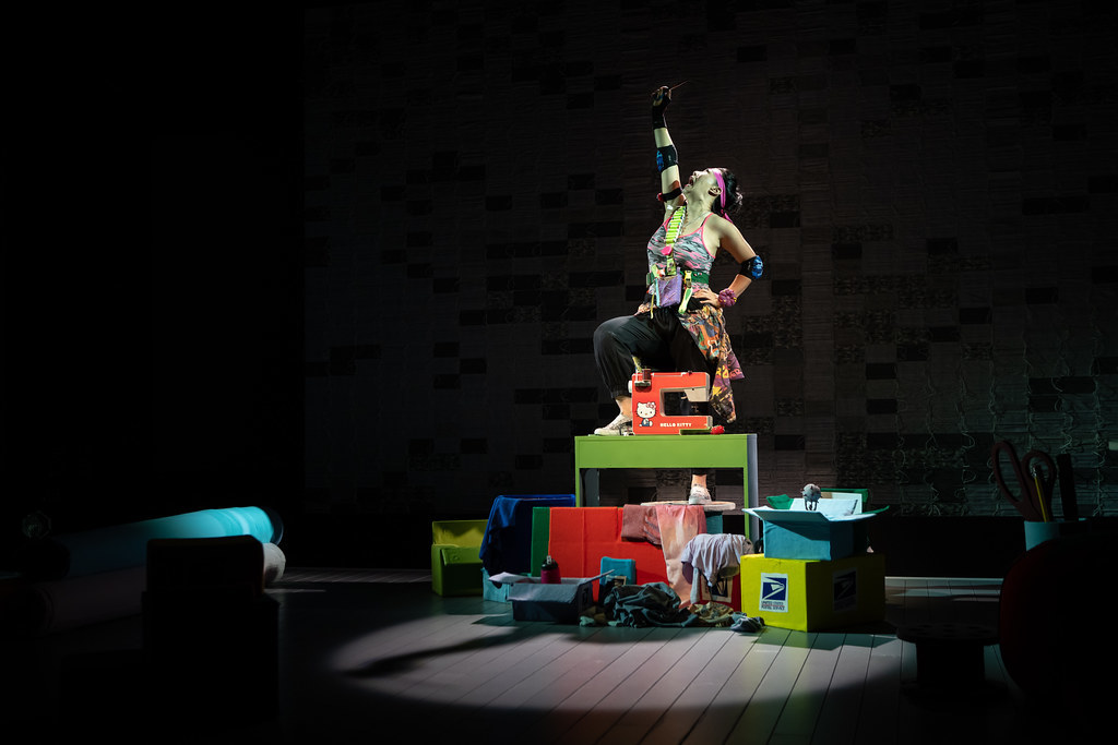 A woman stands triumphantly on a table and proudly holds aloft a pair of scissors.