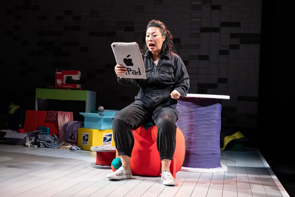 A woman in a black jumpsuit sits on a large tomato pin cushion; she looks at an iPad that fashioned out of felt.