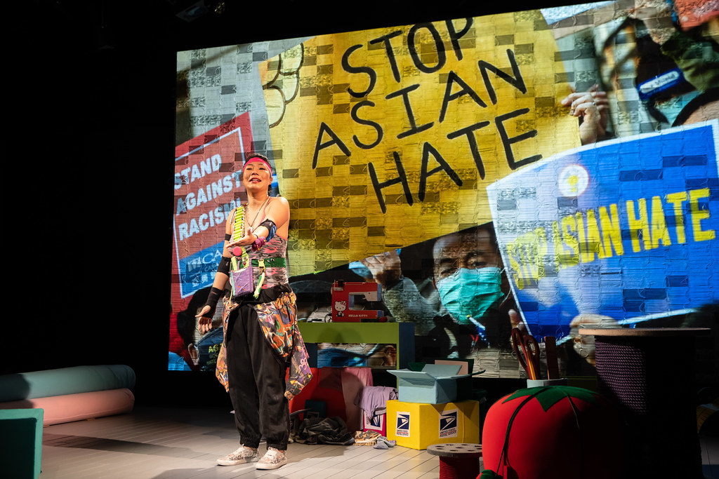 A woman stands in front of a projection of protestors; they hold signs that read Stop Asian Hate and Stand Against Racism. 