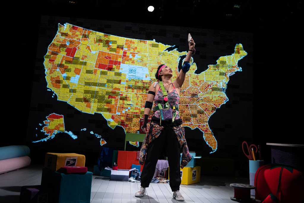 A woman holds up a phone above her head; behind her is a map of the United States representing COVID-19 infection rates.